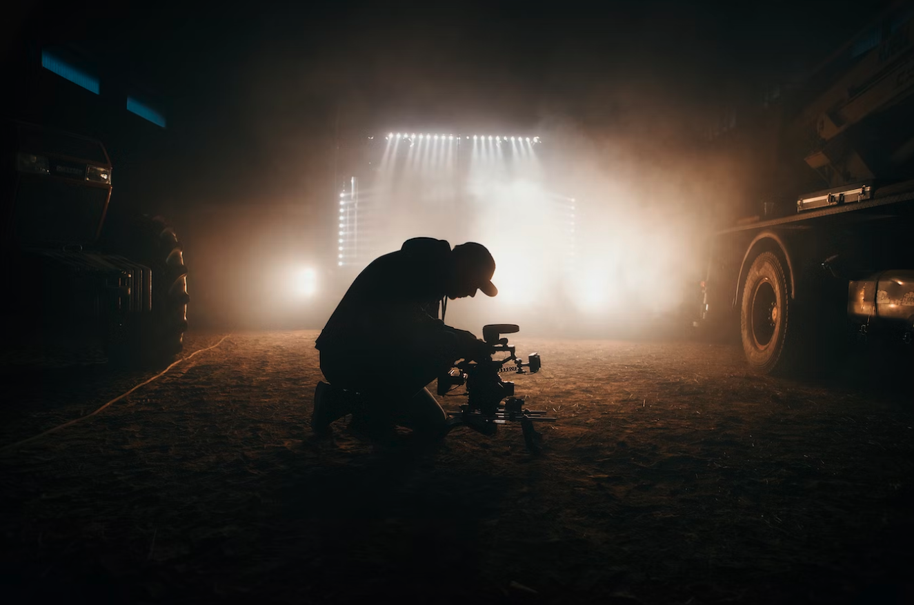 camera operator leaning over a camera on the ground, in the dirt, with two cars on either side and lights shining ahead