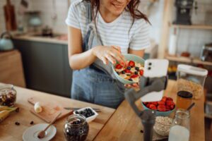 female vlogger holding a bowl with fruit up to a smartphone on a tripod in a home kitchen