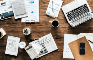 birds eye view of wood desk cluttered with laptop, mug, electronic devices, and paper