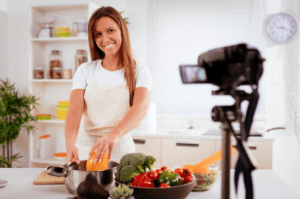 female blogger smiling into a camera on a tripod inside her kitchen while preparing a meal
