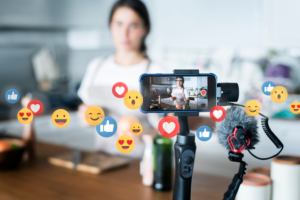 vlogger filming in a kitchen with heart and happy face emojis floating across the picture