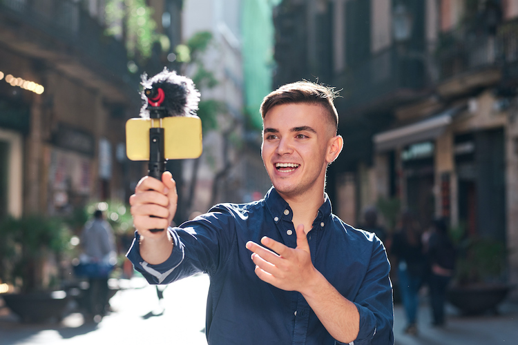 young man in blue shirt recording himself on with a smartphone on a selfie stick on a city street